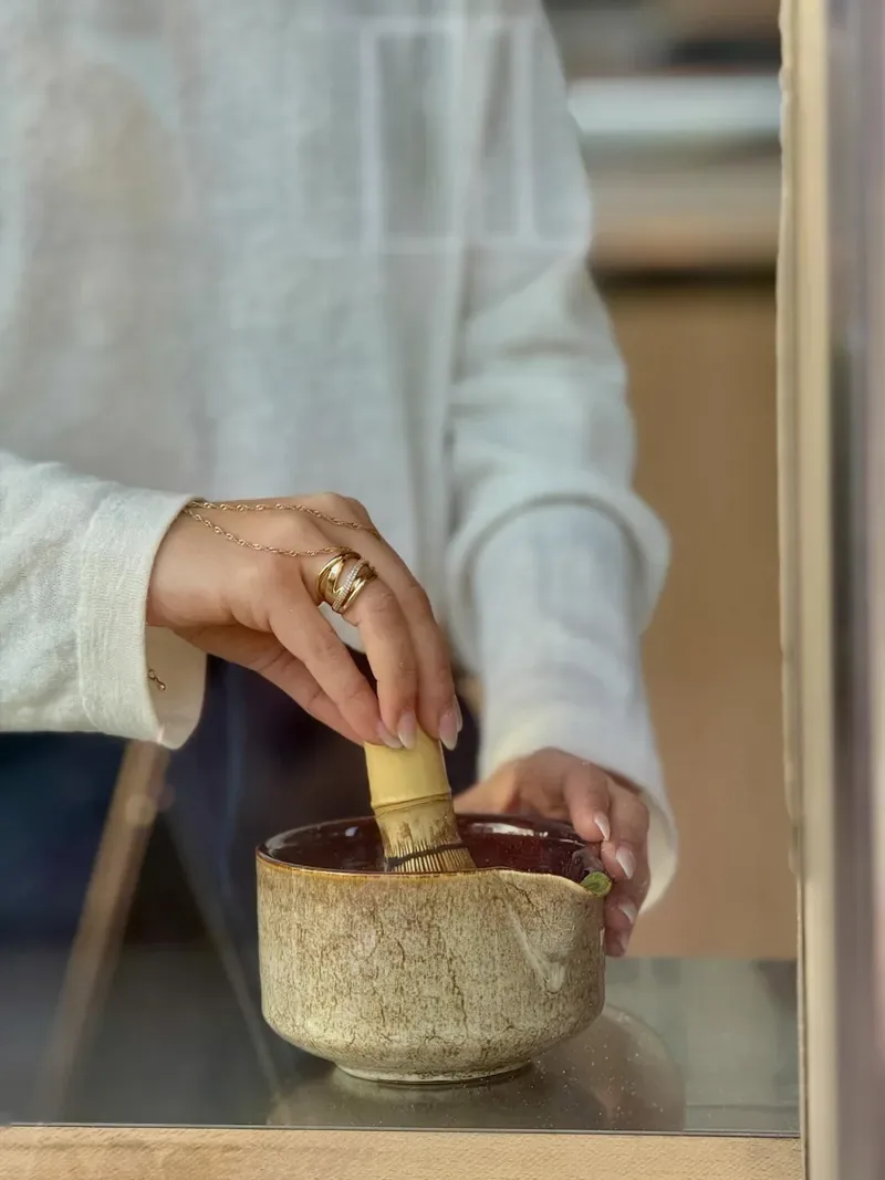 Een hand klopt matcha met een bamboe chasen in een gespikkeld kommetje